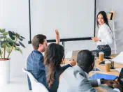 Presenter sits on a conference table. Presentee raises their hand to ask a question.